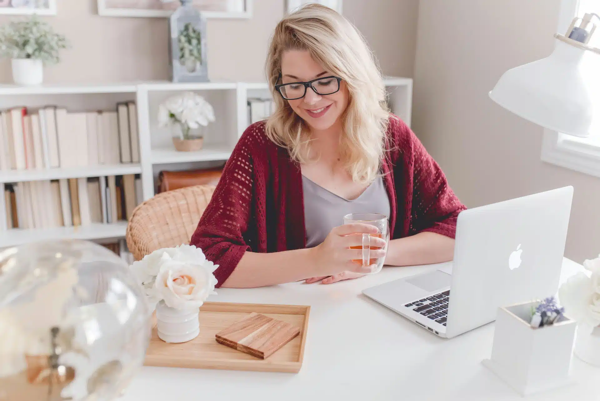 Woman smiling holding glass mug sitting beside table with macbook Woman smiling holding glass mug sitting beside table with macbook